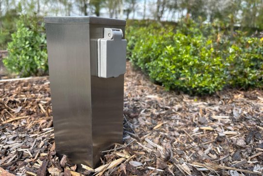 Edelstahlsteckdose im Garten mit Klappdeckel, umgeben von Holzschnitzeln und grüner Vegetation.