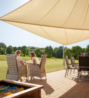 Zwei Personen entspannen sich unter einem Sonnensegel auf einer Terrasse mit Sandkasten und Spielzeug.
