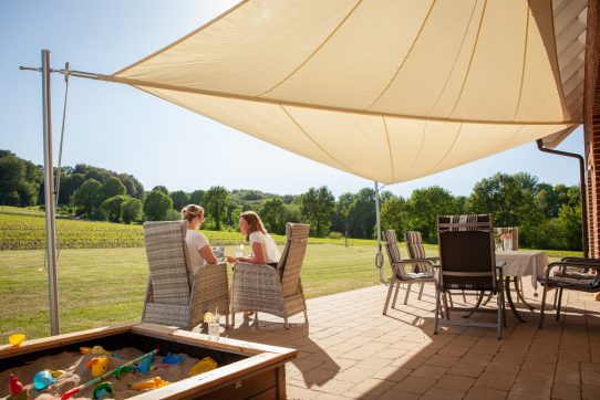 Zwei Personen entspannen sich unter einem Sonnensegel auf einer Terrasse mit Sandkasten und Spielzeug.