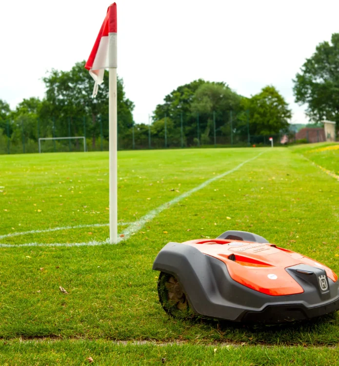 Ein automatischer Rasenmäher mäht den grünen Rasen eines Sportplatzes neben einer rot-weißen Eckfahne.