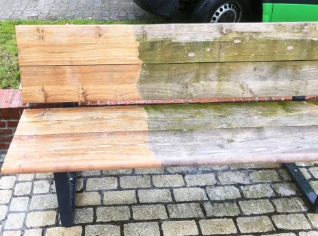Eine Holzbank, halb gereinigt, halb verwittert, steht auf Pflastersteinen mit einem Auto im Hintergrund.