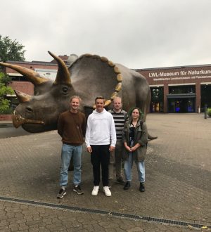 Vier Personen posieren vor einer realistischen Triceratops-Statue vor dem LWL-Museum für Naturkunde.